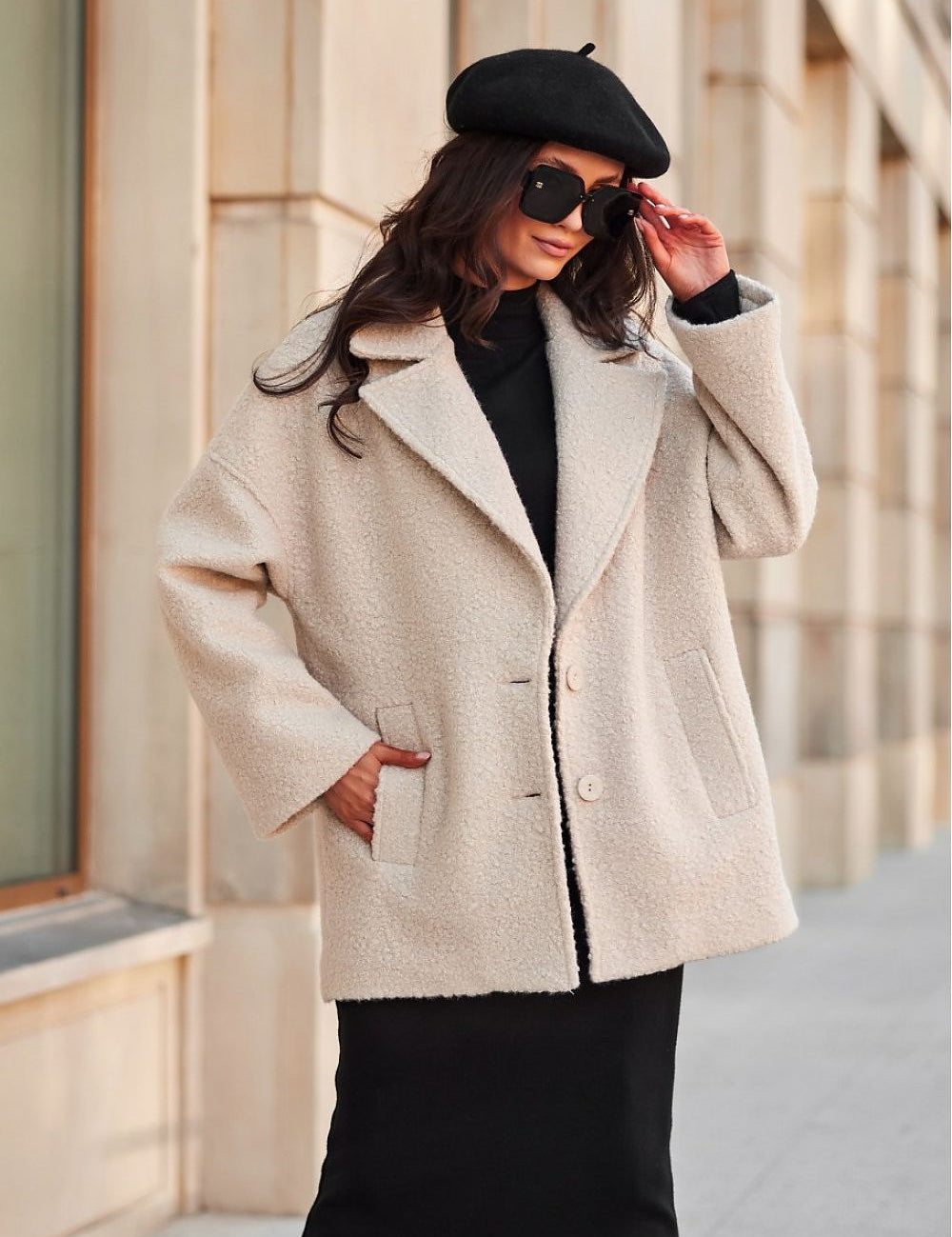 Stylish woman in a beige wool coat and black beret with sunglasses, standing on a city sidewalk.