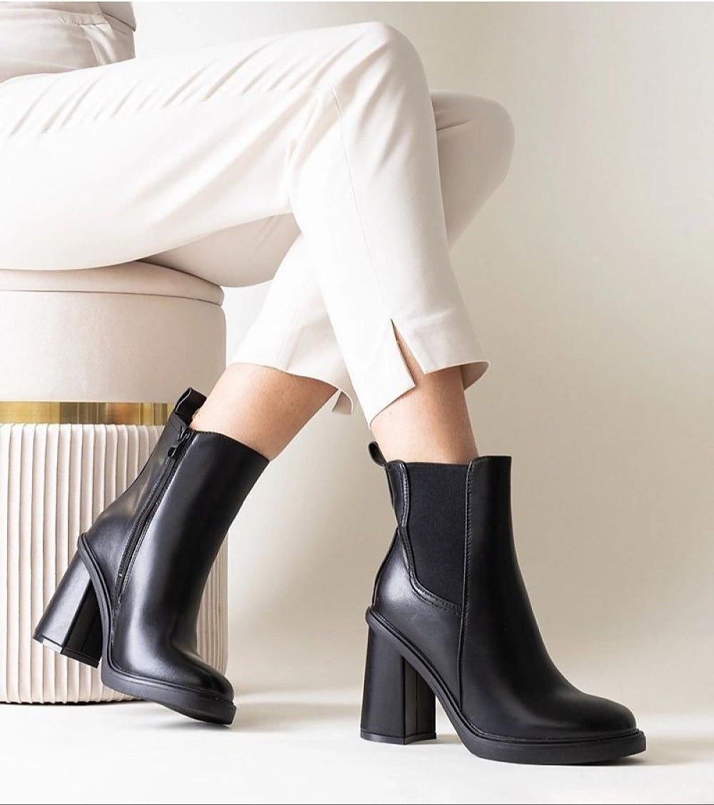 Black stiletto zip-up boots with eco leather, high heel, and side zipper, worn with white pants, on minimal background.