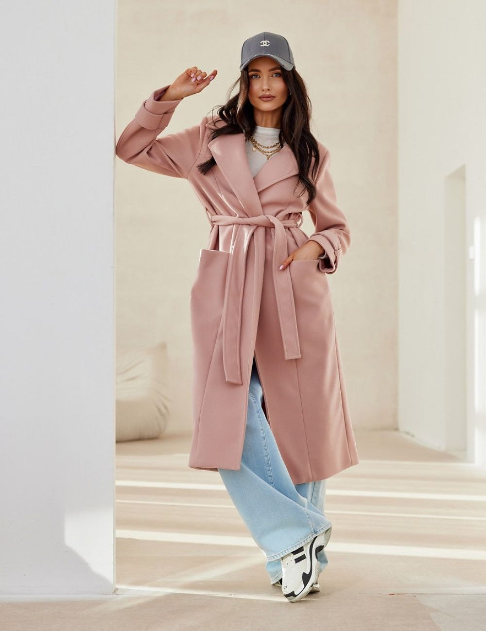 Woman wearing a Loravinne belted long-sleeve lined coat in light pink, paired with blue jeans and sneakers, standing in a bright room.