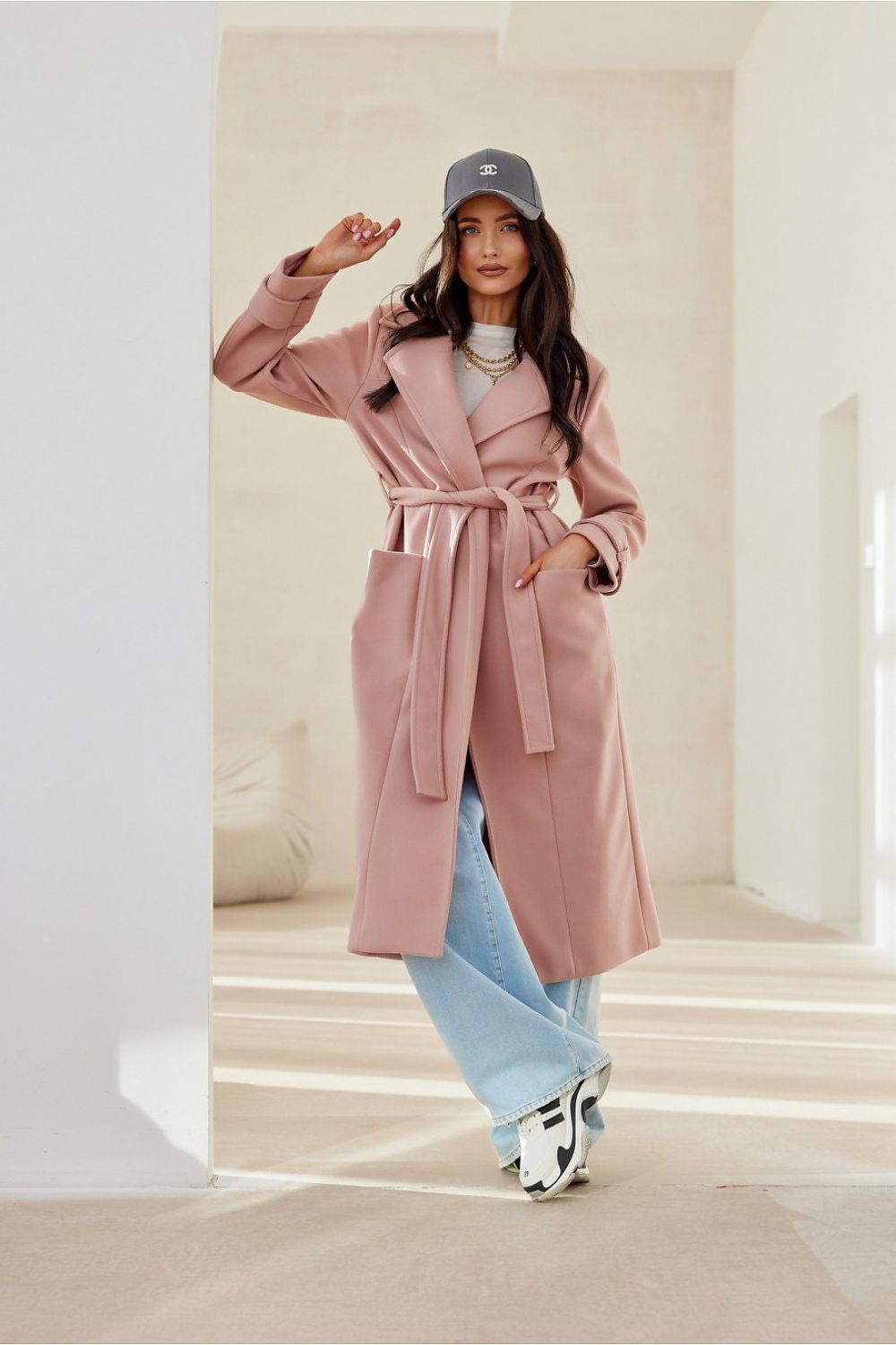 Woman wearing a Loravinne belted long-sleeve lined coat in light pink, paired with blue jeans and sneakers, standing in a bright room.