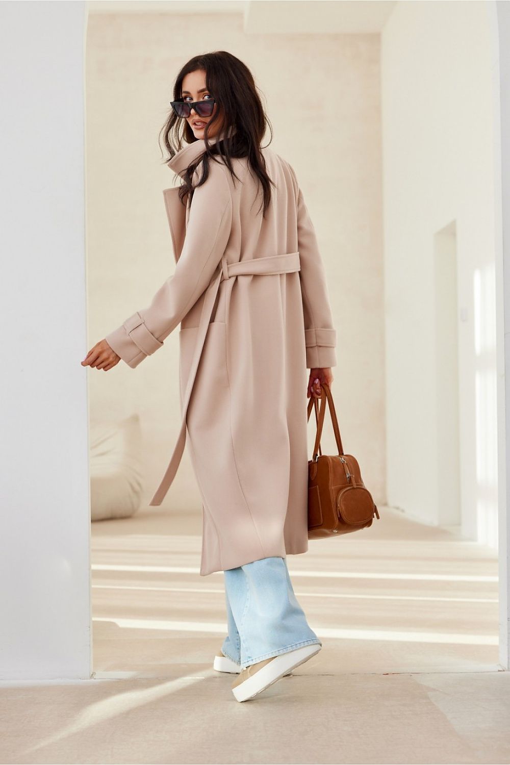 Woman wearing Loravinne Belted Long-Sleeve Coat with brown handbag and sunglasses, showing back view in bright indoor setting.