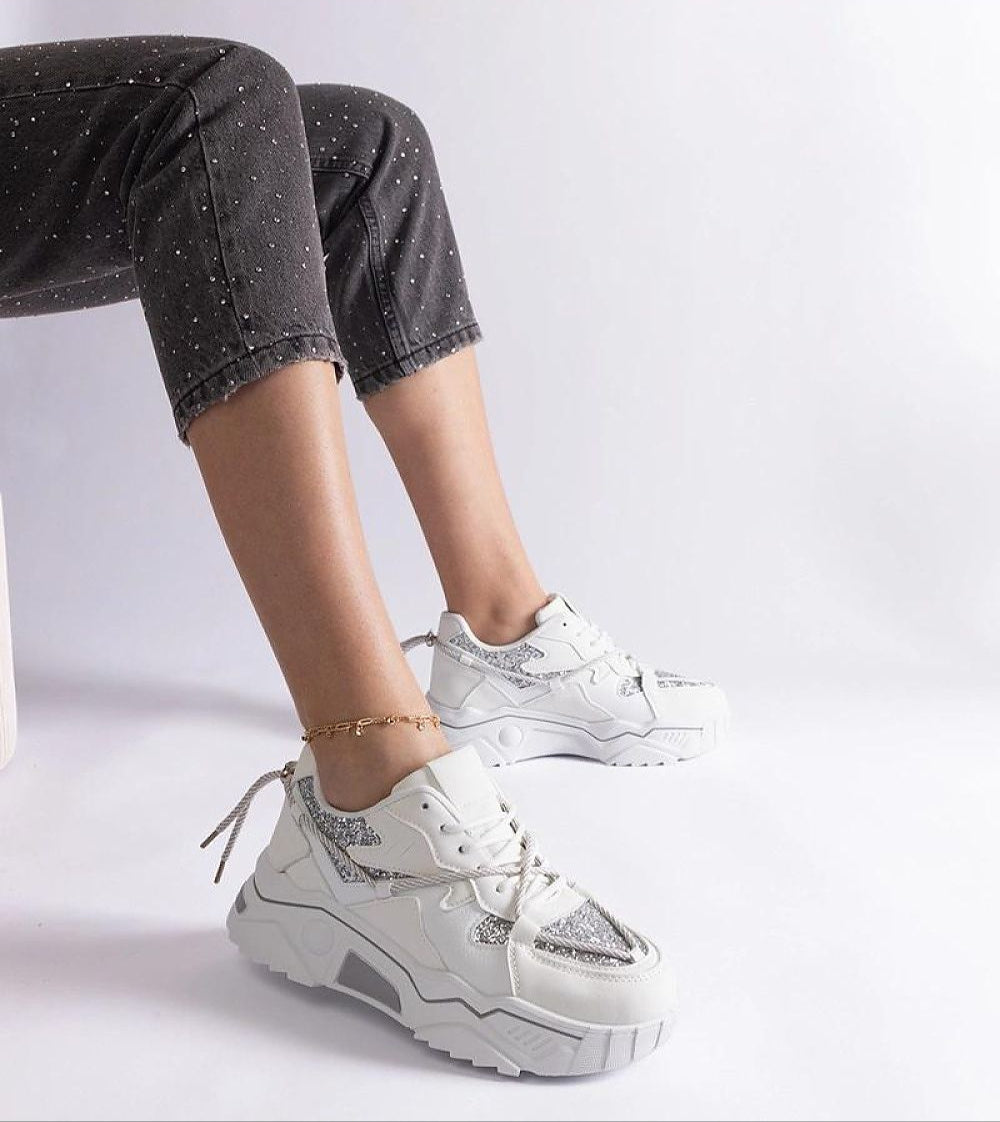 Person wearing white and silver sneakers with black jeans, ankle bracelet, seated on a white chair.