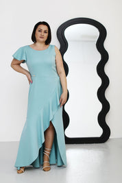 Plus size model wearing a blue one-shoulder cocktail dress with ruffle hem, standing near a wavy mirror.