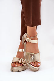 Gold buckled Talmera platform sandals with eco leather finish and flat sole, styled with brown trousers for a chic, minimalist look.