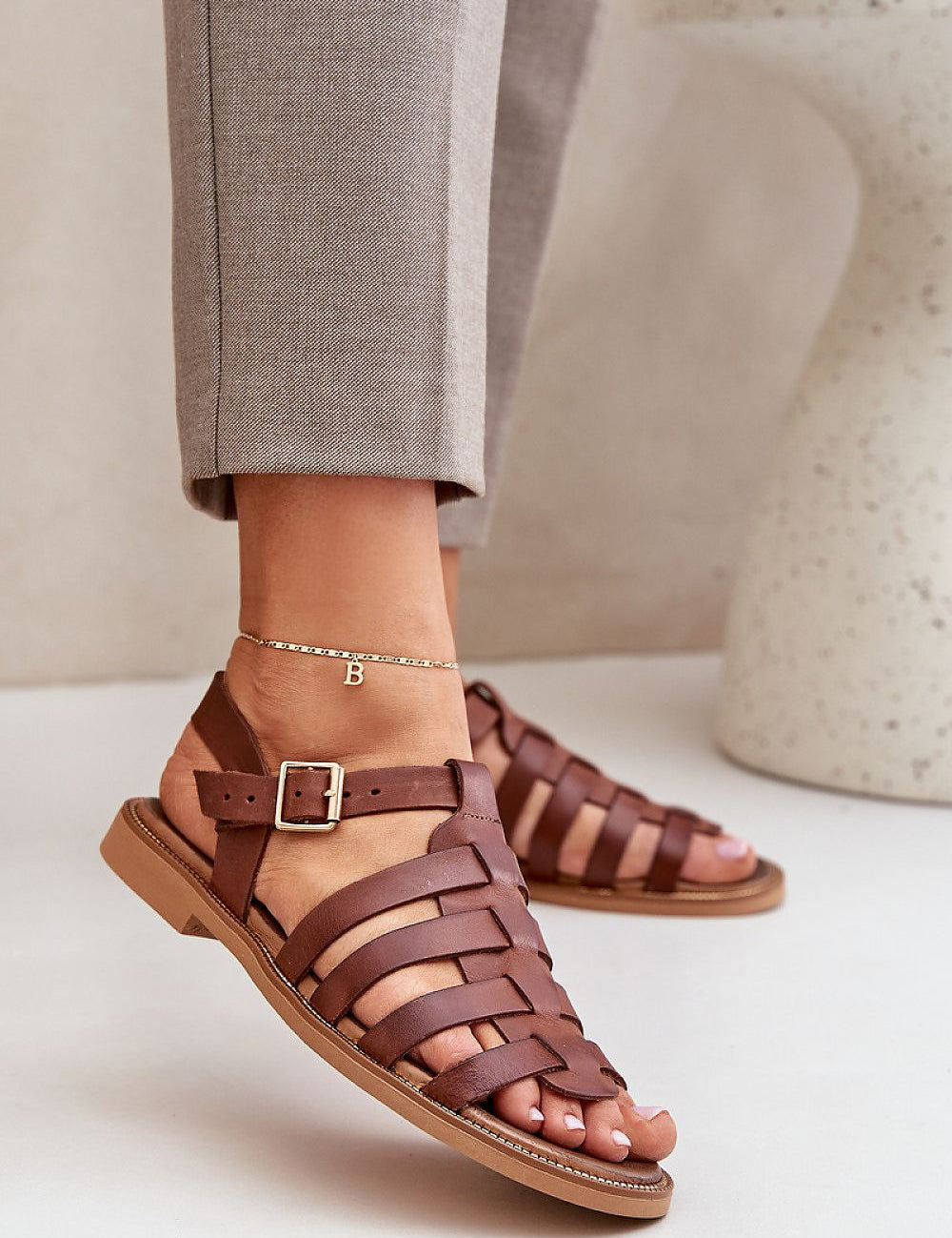 Woman wearing Celira Flat Leather Sandals with ankle buckle in brown, showcasing a sleek, elegant design perfect for versatile wear.