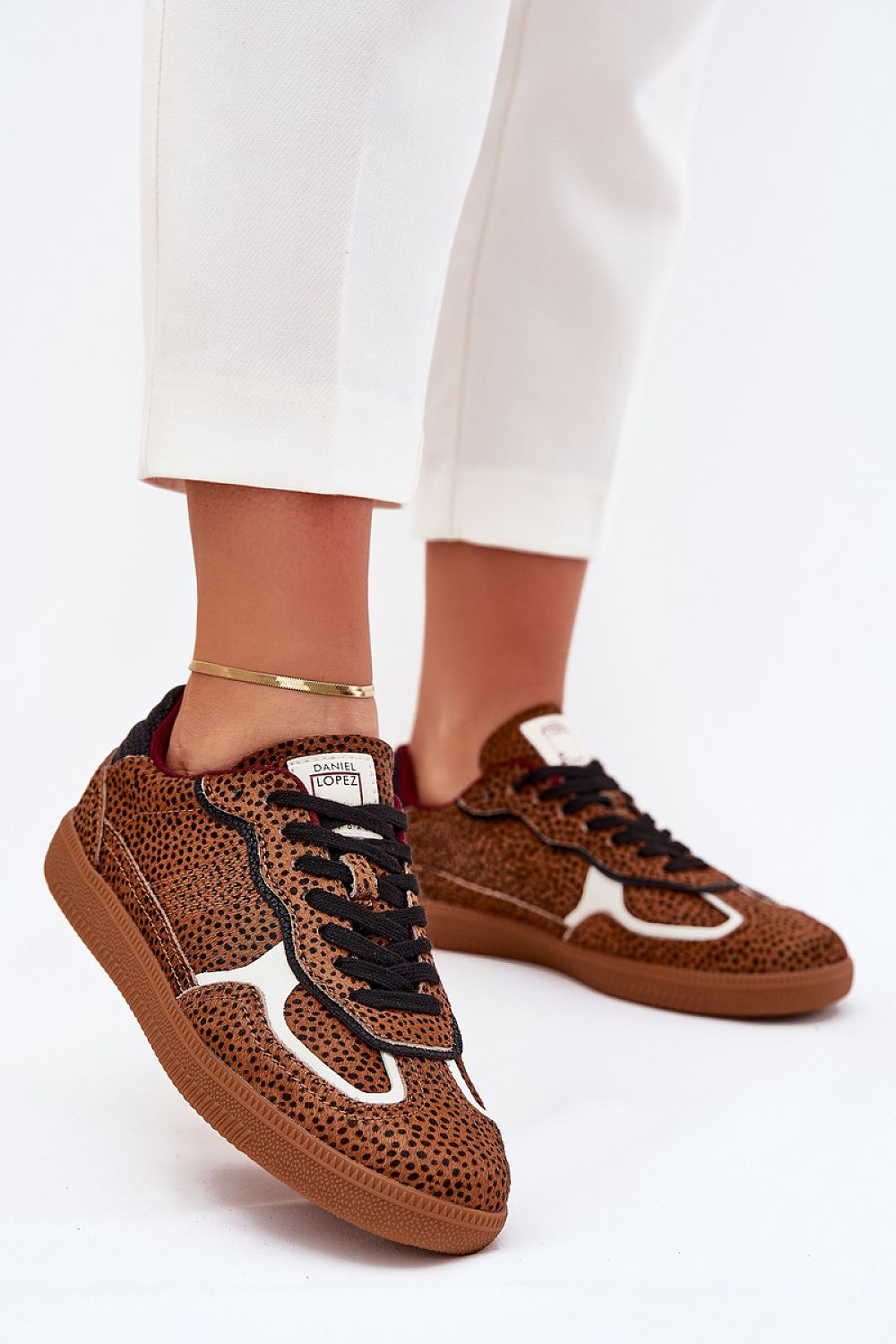 Close-up of a person wearing brown speckled sneakers with black laces and white accents, paired with white pants.