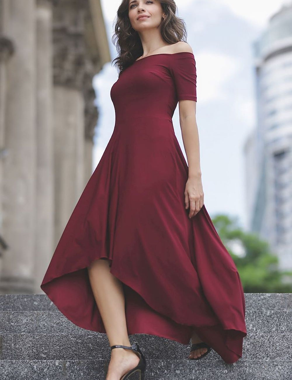 Woman wearing Aymera One-Shoulder Cocktail Dress in burgundy, standing on steps against city backdrop.