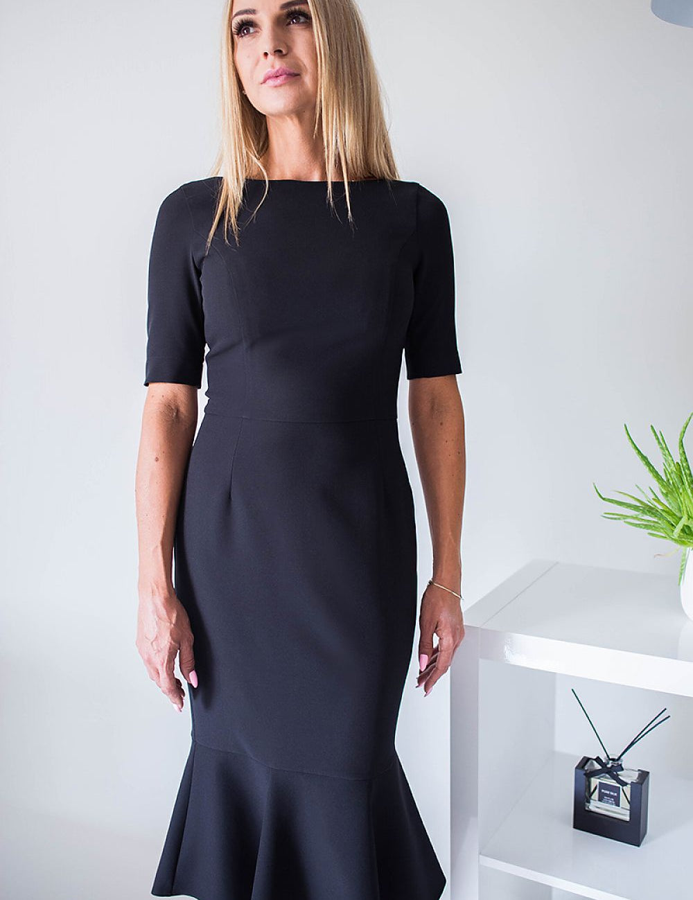 Woman wearing a black plus size Rivoria zipped cocktail dress with elbow-length sleeves and frill hem, standing in a modern room.