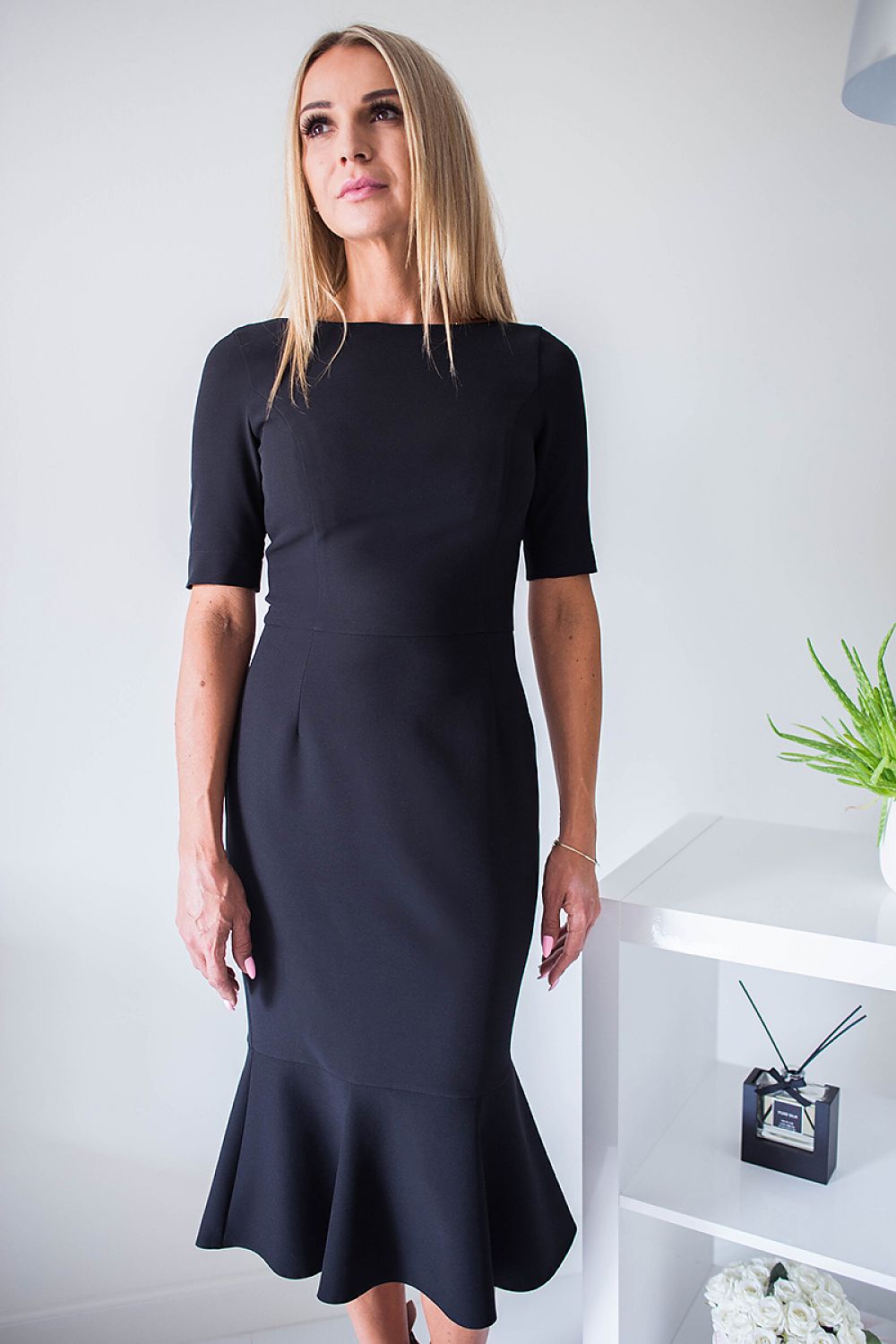 Woman wearing a black plus size Rivoria zipped cocktail dress with elbow-length sleeves and frill hem, standing in a modern room.