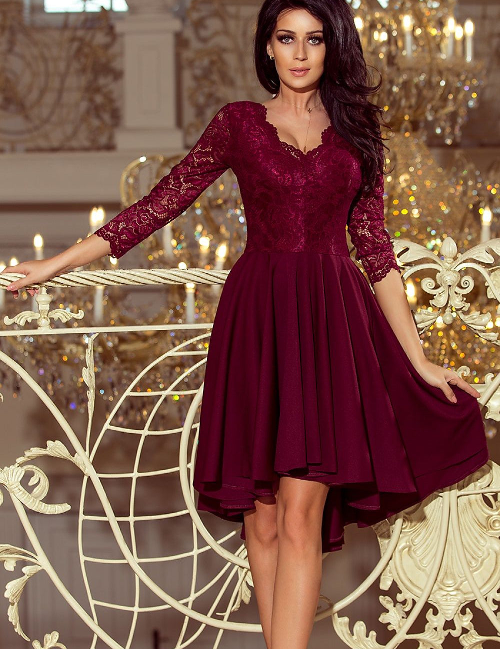 Woman wearing Nivelle Burgundy Lace High-Low Dress with lace bodice and asymmetrical hem, standing elegantly on ornate balcony.