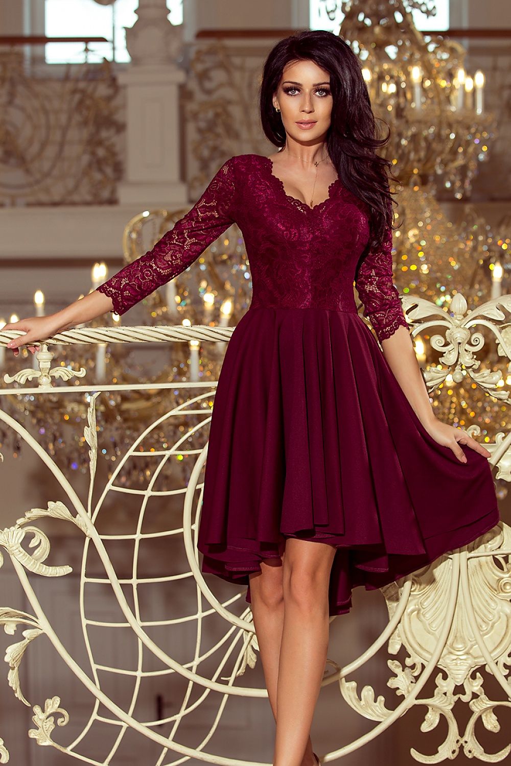Woman wearing Nivelle Burgundy Lace High-Low Dress with lace bodice and asymmetrical hem, standing elegantly on ornate balcony.