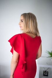 Woman wearing red dress with ruffle sleeves, standing sideways in a room with light gray walls and a small plant in the background.