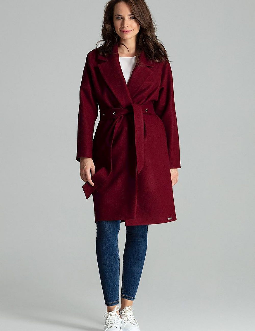 Woman wearing Carivelle Maroon Belted Coat with Wide Lapels, featuring metal eyelets and side-seam pockets, styled with jeans and sneakers.