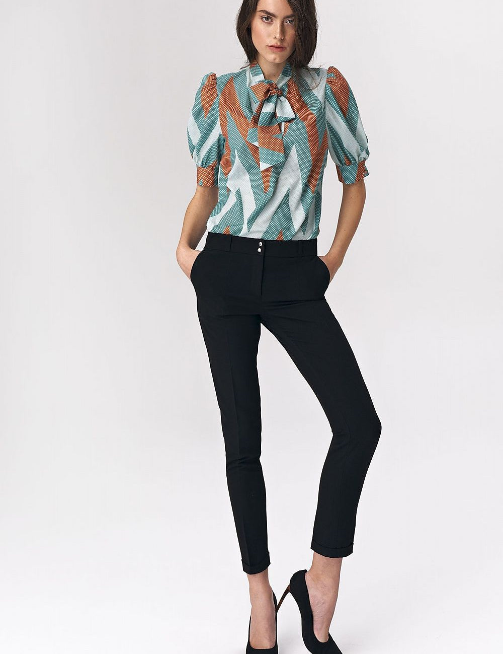 Woman wearing Myressia Zigzag Tie Blouse in turquoise with zigzag pattern, paired with black trousers and heels, hands in pockets.