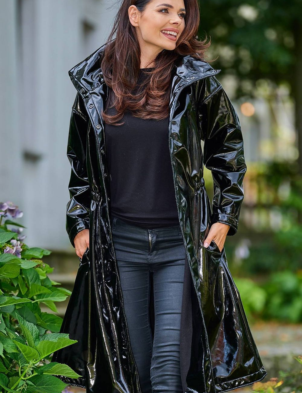Woman wearing Marveena knee-length black vinyl raincoat with high collar and flared hem, standing outdoors.