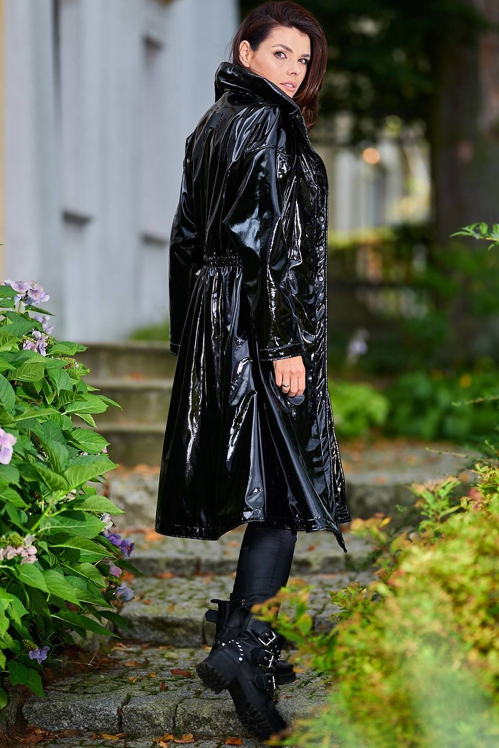 Woman wearing black Marveena knee-length vinyl raincoat on garden path