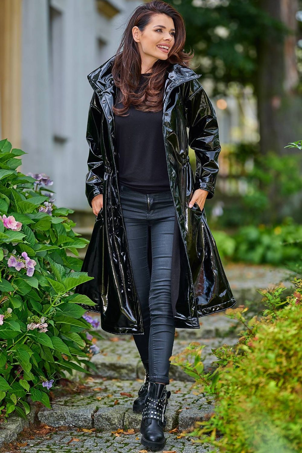 Woman wearing Marveena knee-length black vinyl raincoat with high collar, sleek profile, walking in garden