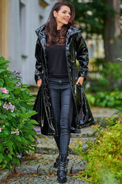 Woman wearing Marveena knee-length black vinyl raincoat with high collar, sleek profile, walking in garden