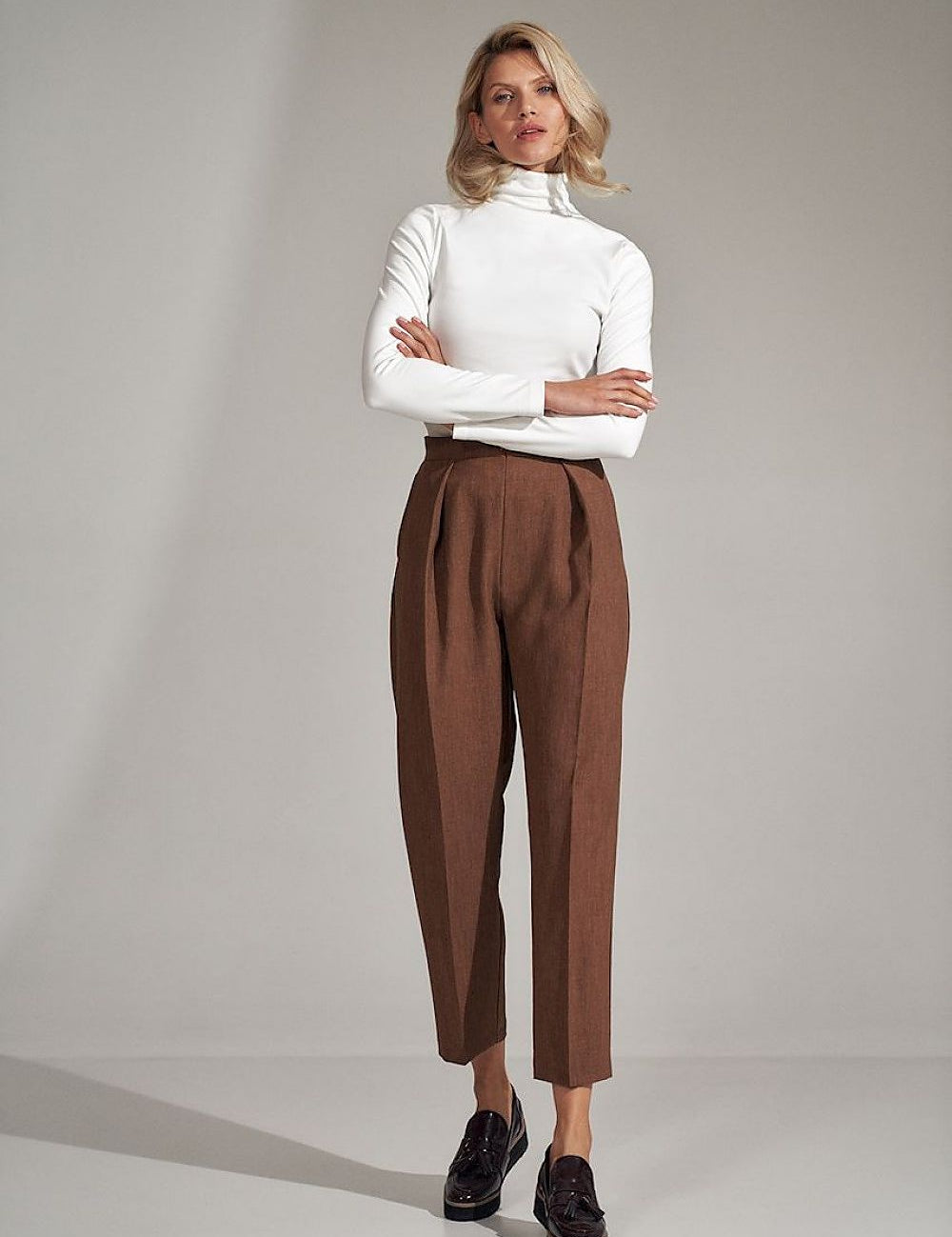 Woman wearing Mireval Turtleneck Blouse in soft ecru, paired with brown high-waisted trousers, showcasing a classic and elegant style.