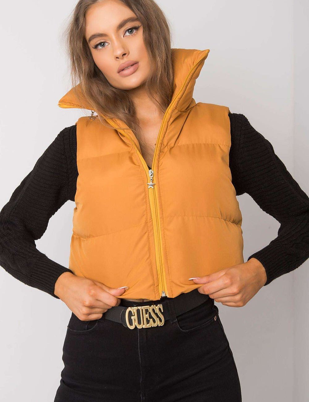 Woman wearing an orange zip vest with high collar and black sleeves, featuring a Guess belt buckle.
