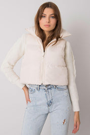 Woman wearing white sleeveless puffer vest and jeans, standing against a gray background.