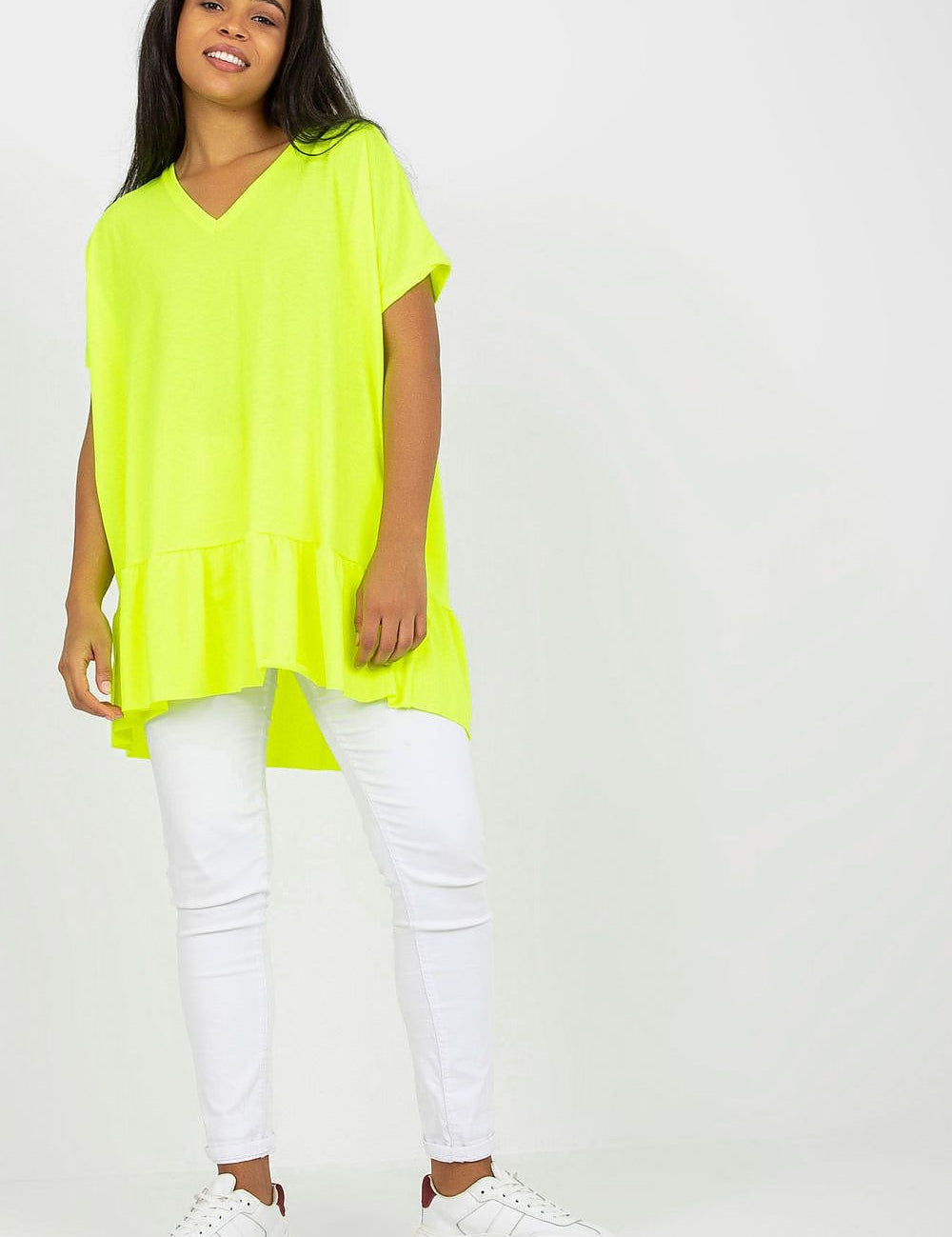 Model wearing a lime green plus size tunic with frilled hem and heart-shaped neckline, paired with white leggings and sneakers.