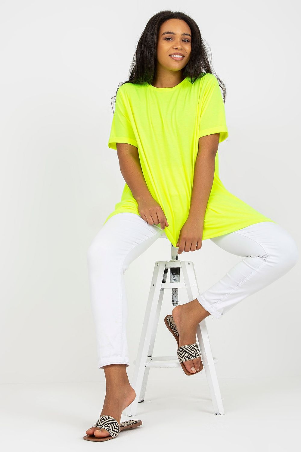 Woman wearing a neon yellow tunic with white pants, seated on a stool, demonstrating a relaxed and casual style.