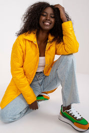 Woman modeling Cerenya quilted jacket with extended back, wearing casual jeans and sneakers, showcasing quilt design and vibrant color.