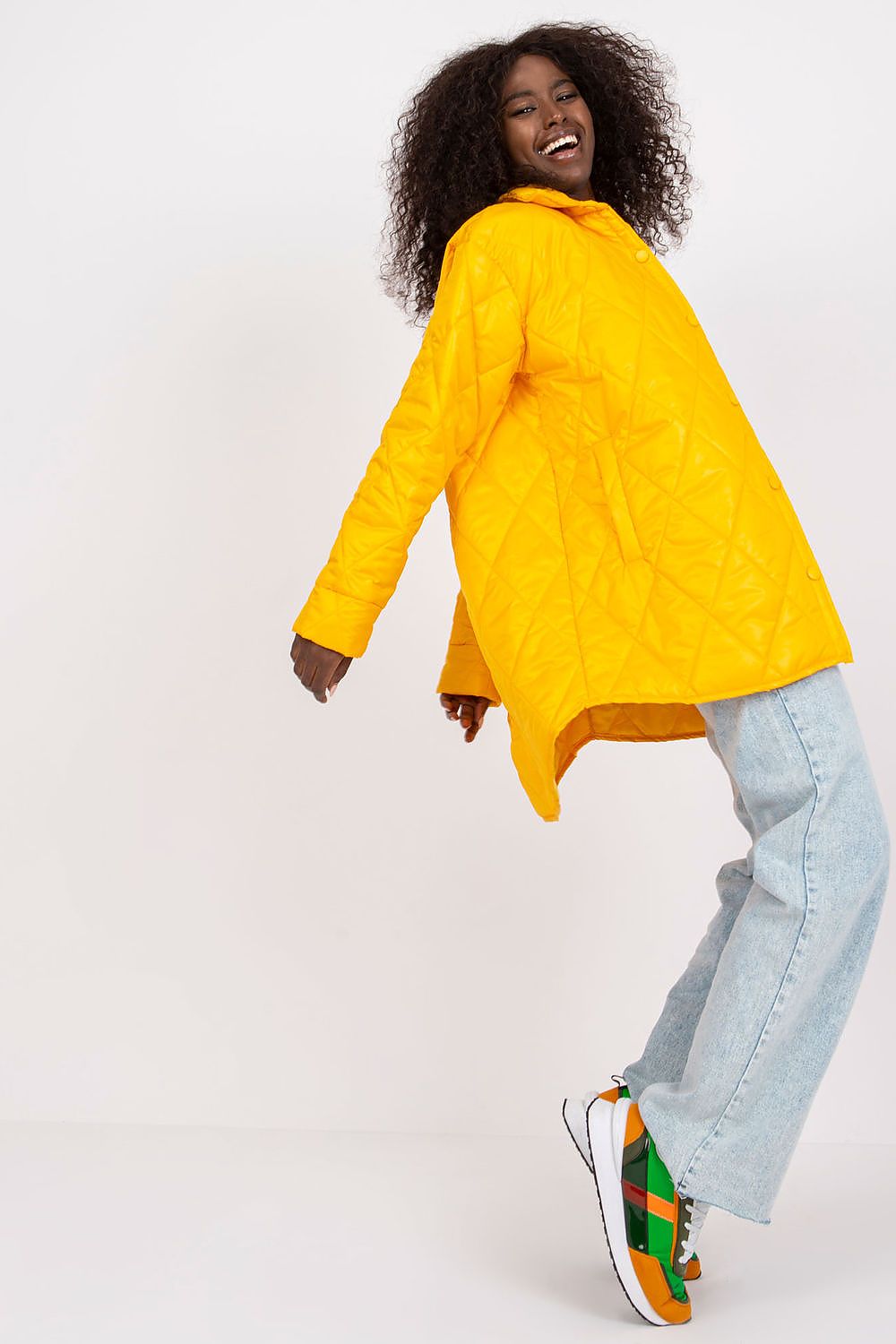 Woman wearing Cerenya Quilted Jacket in yellow with extended back, perfect for transitional seasons, paired with jeans and colorful sneakers.