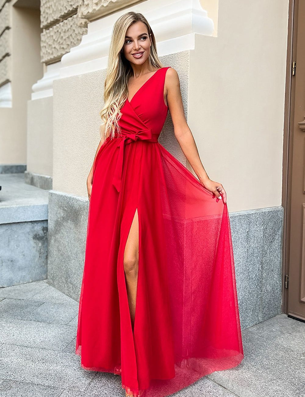 Woman wearing a red evening dress with a V-neck and high slit, standing outdoors against a light-colored building wall.