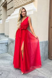 Woman wearing a red evening dress with a V-neck and high slit, standing outdoors against a light-colored building wall.