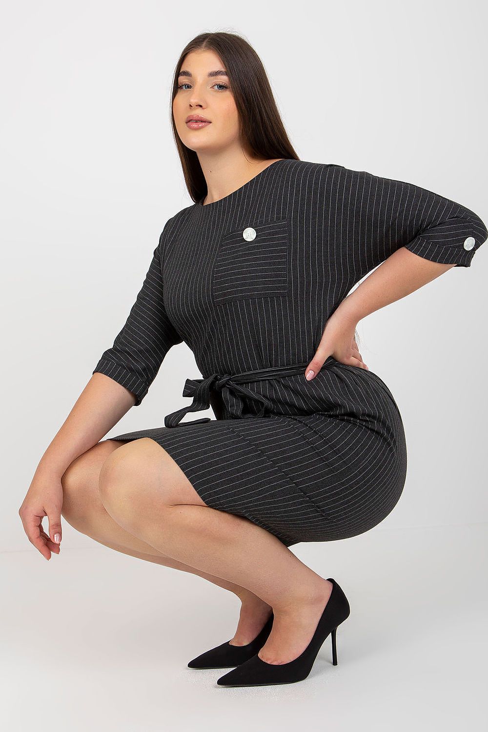Model wearing Velmira Plus Size Striped Pencil Dress, kneeling, showcasing 3/4 sleeves, round neckline, and tie belt for flattering fit.