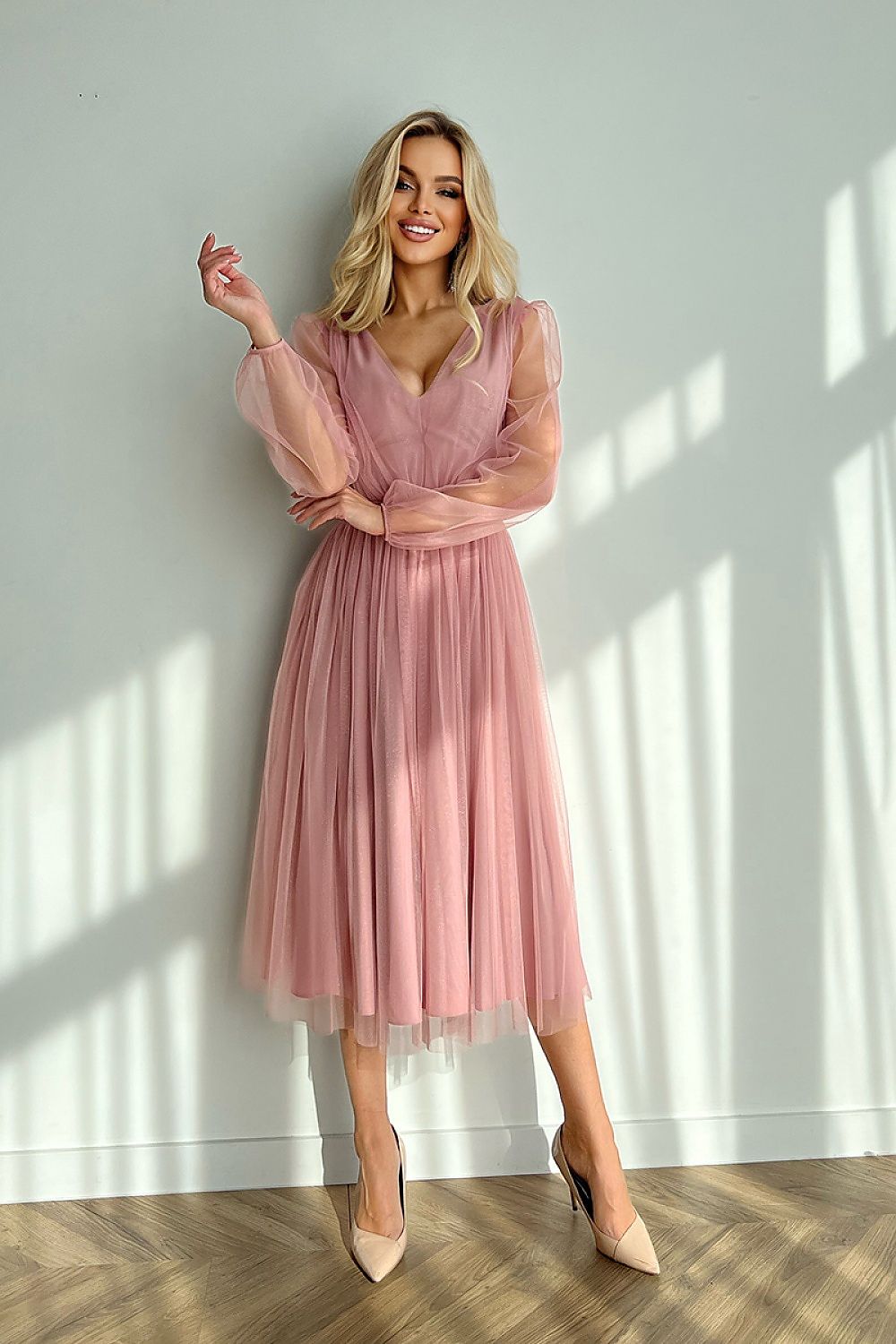 Blonde woman wearing a pink mesh-layered midi dress with sheer sleeves and brocade detailing, standing against a sunlit wall.