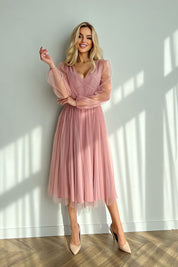 Blonde woman wearing a pink mesh-layered midi dress with sheer sleeves and brocade detailing, standing against a sunlit wall.
