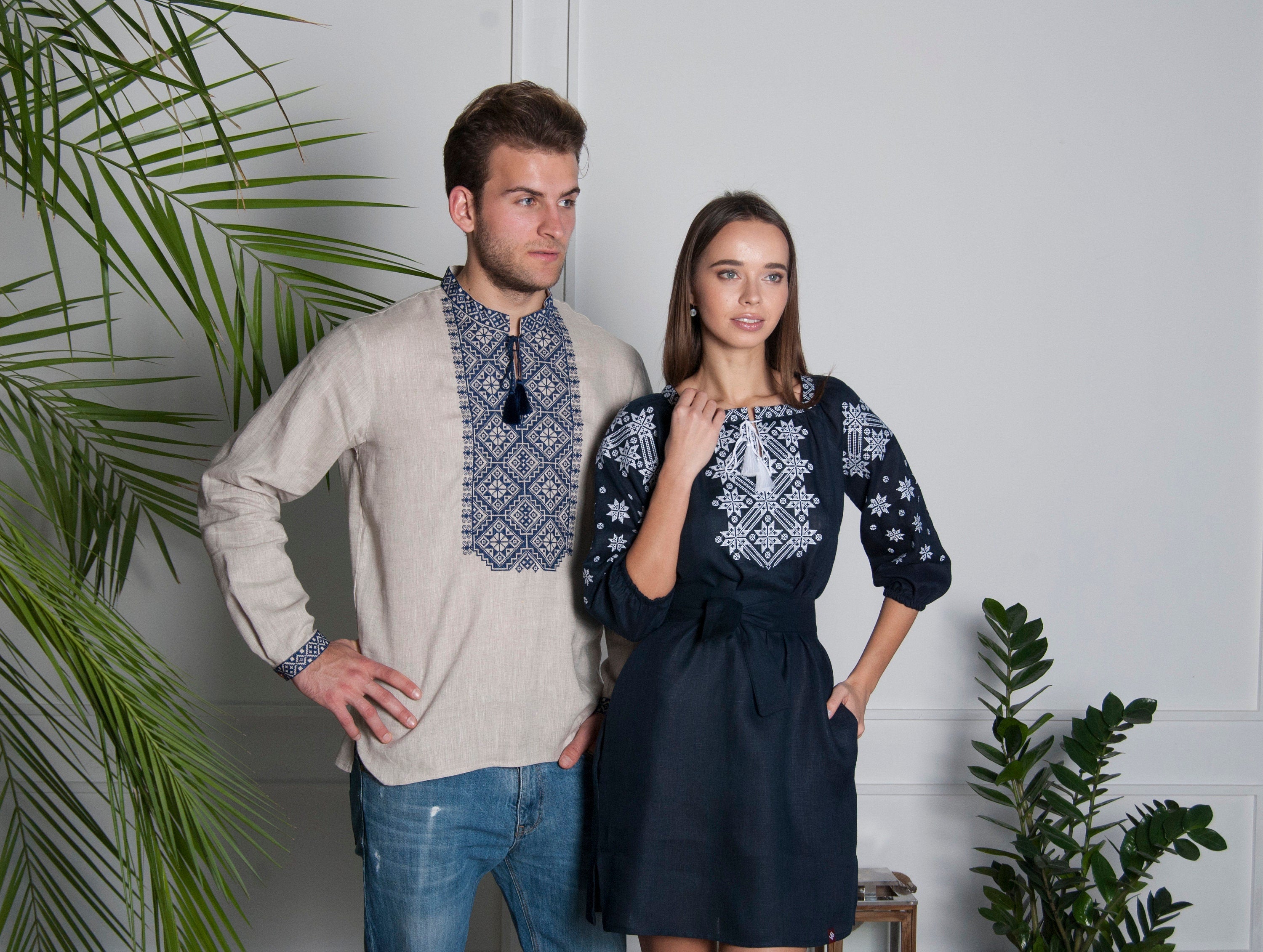 Man in embroidered beige shirt and woman in dark blue linen dress with Alatyr symbol, traditional Ukrainian vyshyvanka attire.