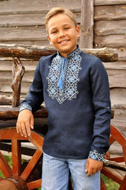 Boy wearing dark blue linen Ukrainian Vyshyvanka shirt with Alatyr embroidery, standing outdoors by wooden wheel, smiling.