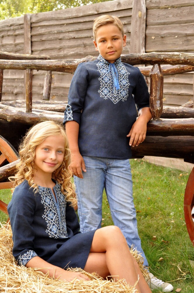 Boy and girl wearing dark blue Ukrainian vyshyvanka shirts with Alatyr symbol embroidery, sitting on a haystack outdoors.