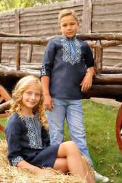Boy and girl wearing dark blue Ukrainian vyshyvanka shirts with Alatyr symbol embroidery, sitting on a haystack outdoors.