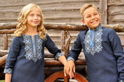 Two children in dark blue embroidered Ukrainian vyshyvanka shirts with Alatyr symbols, standing by a wooden fence.