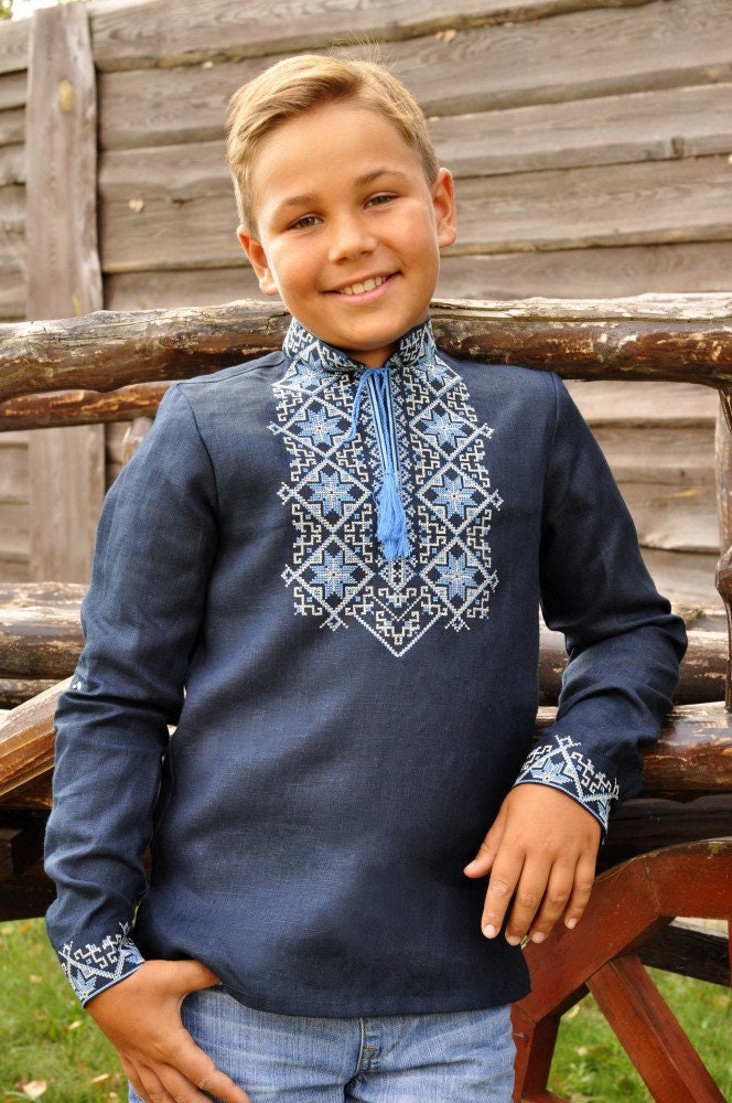 Boy wearing dark blue linen embroidered Ukrainian Vyshyvanka shirt with Alatyr symbol, standing outdoors.