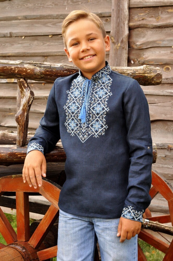 Boy wearing dark blue Ukrainian linen shirt with blue embroidery and Alatyr symbol, standing by wooden wagon.