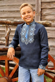 Boy wearing dark blue Ukrainian linen shirt with blue embroidery and Alatyr symbol, standing by wooden wagon.