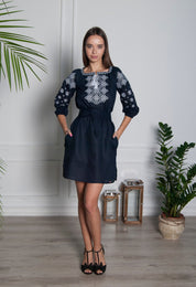 Woman wearing dark blue linen embroidered dress with Ukrainian Alatyr symbol in a room decorated with plants.