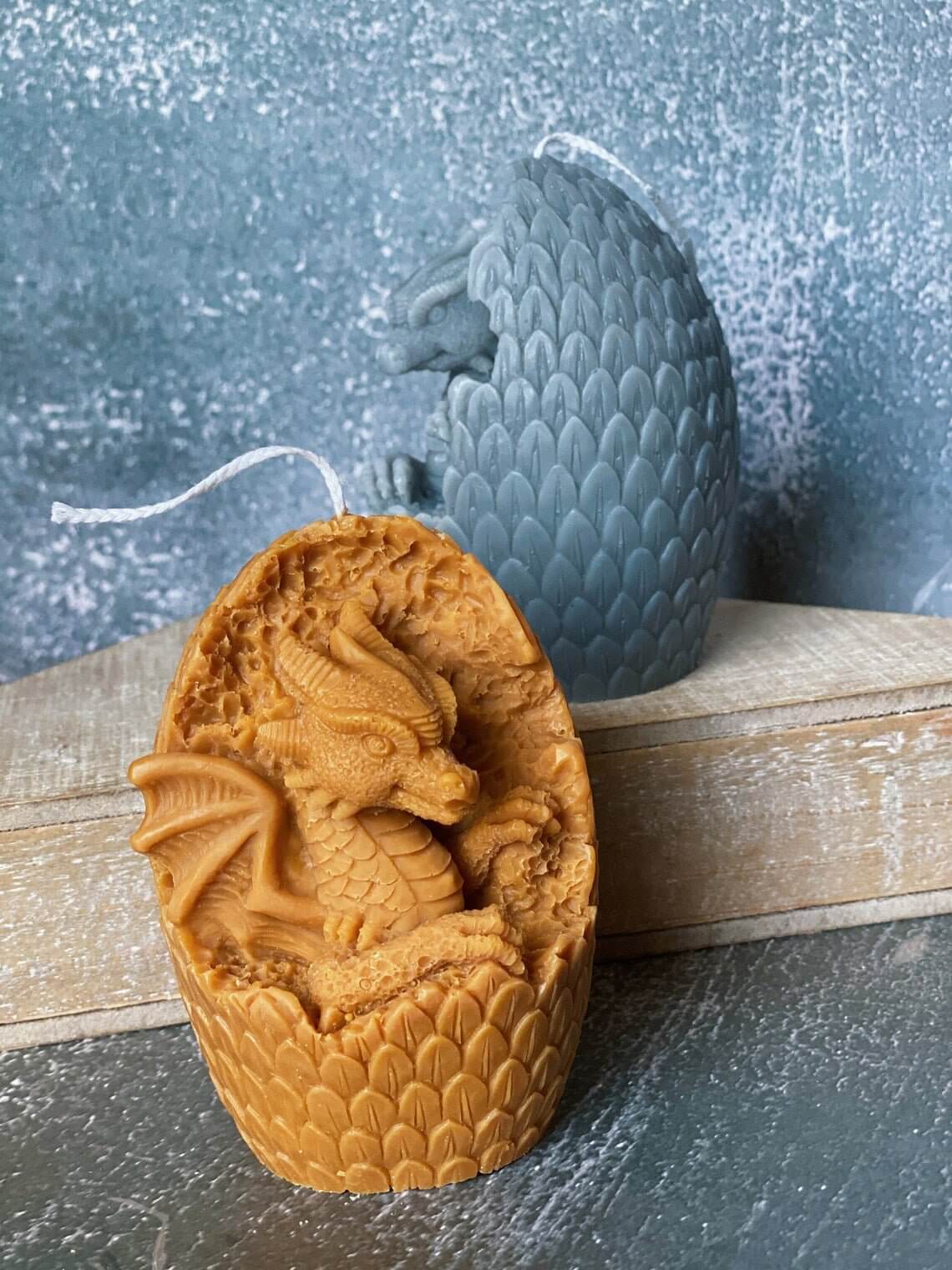 Two handmade dragon's egg candles, one carved open showing a dragon, crafted from natural soy wax with textured scales in various colors.