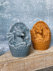 Two handmade dragon's egg candles with detailed dragon designs, crafted from soy wax in blue and amber shades, set on a wooden plank.