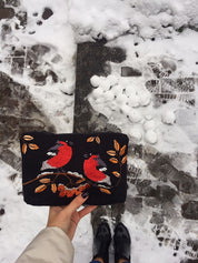 Person holding embroidered makeup bag with Ukrainian pattern, featuring birds and leaves, over snow-covered ground.