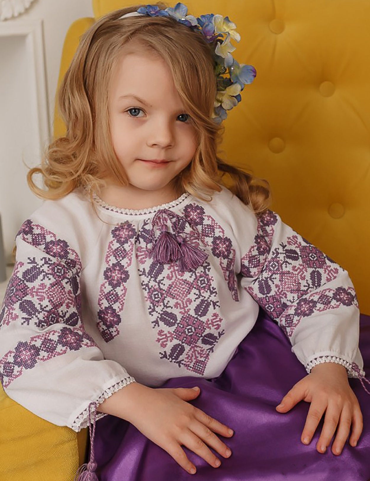 Girl wearing embroidered Ukrainian blouse with floral patterns, seated on yellow chair.