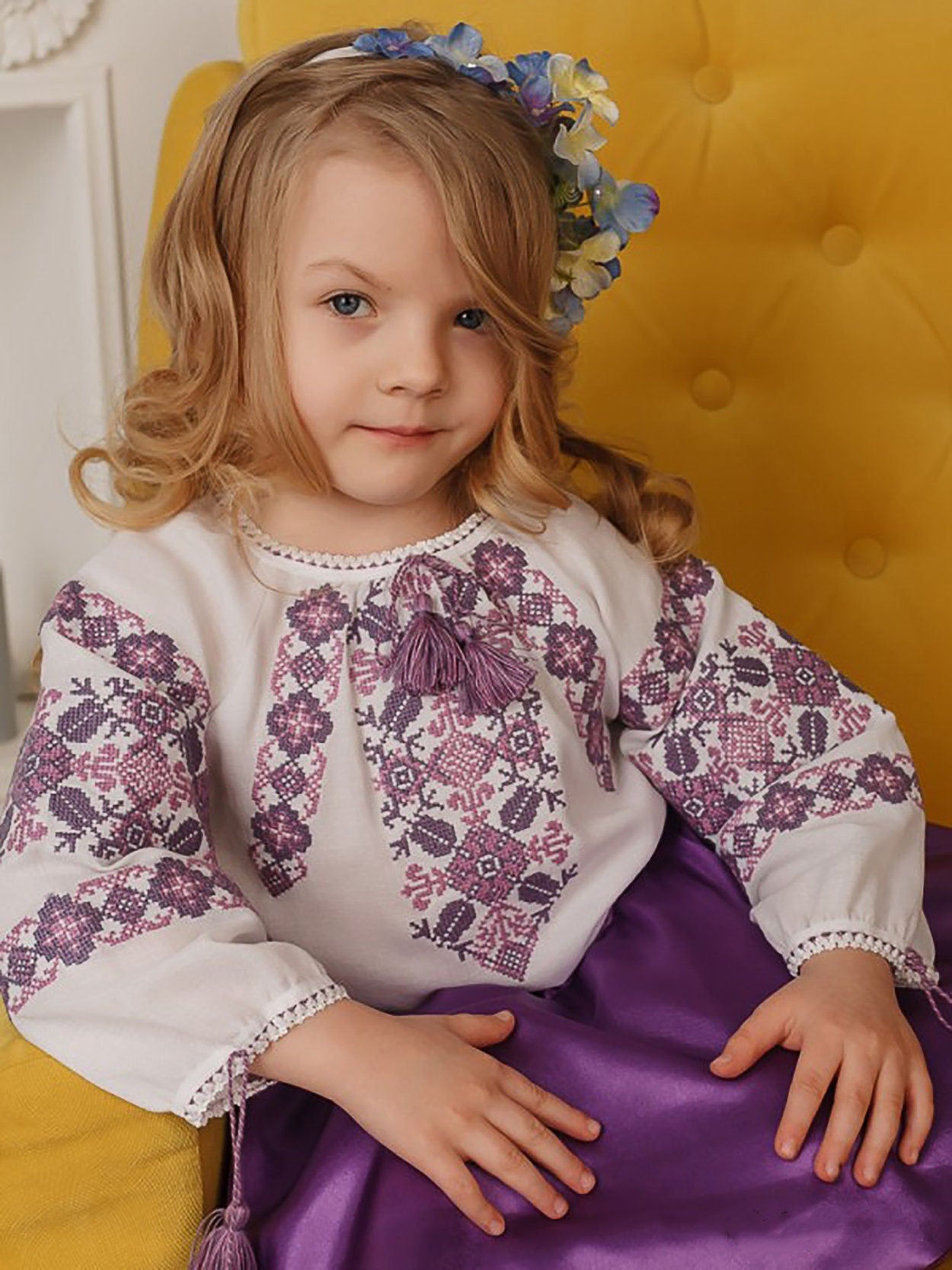 Girl wearing embroidered Ukrainian blouse with floral patterns, seated on yellow chair.