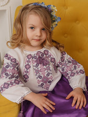 Girl wearing embroidered Ukrainian blouse with floral patterns, seated on yellow chair.