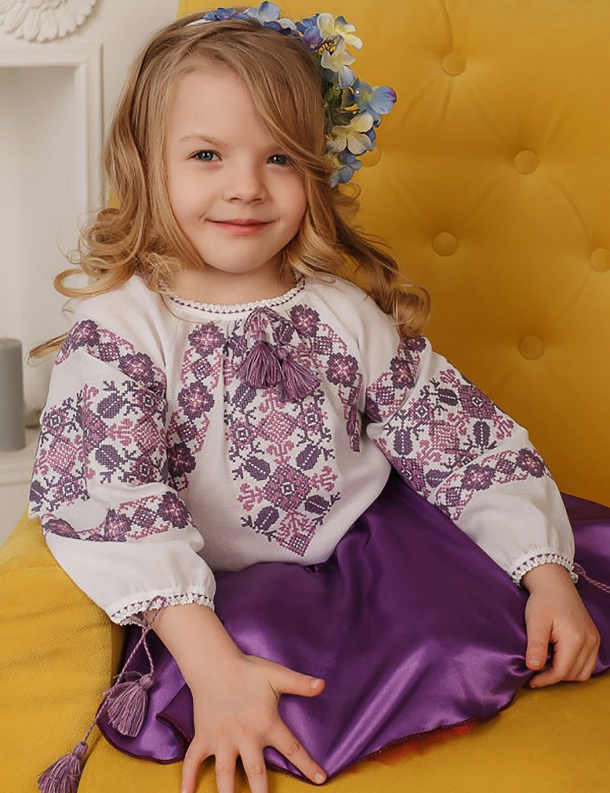 Young girl in traditional embroidered Ukrainian blouse with cross-stitch patterns sitting on yellow chair.